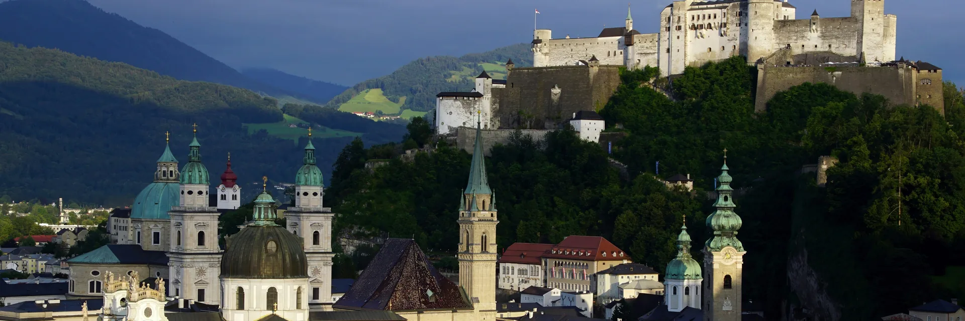 Hohensalzburg Fortress's white walls above Salzburg's old town domes and rooftops, seen across the city at dusk